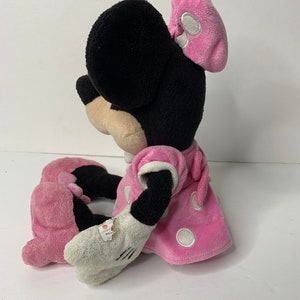May include: A pink plush Minnie Mouse toy with white polka dots on her dress and bow. She is sitting with her legs crossed and her arms resting on her lap.