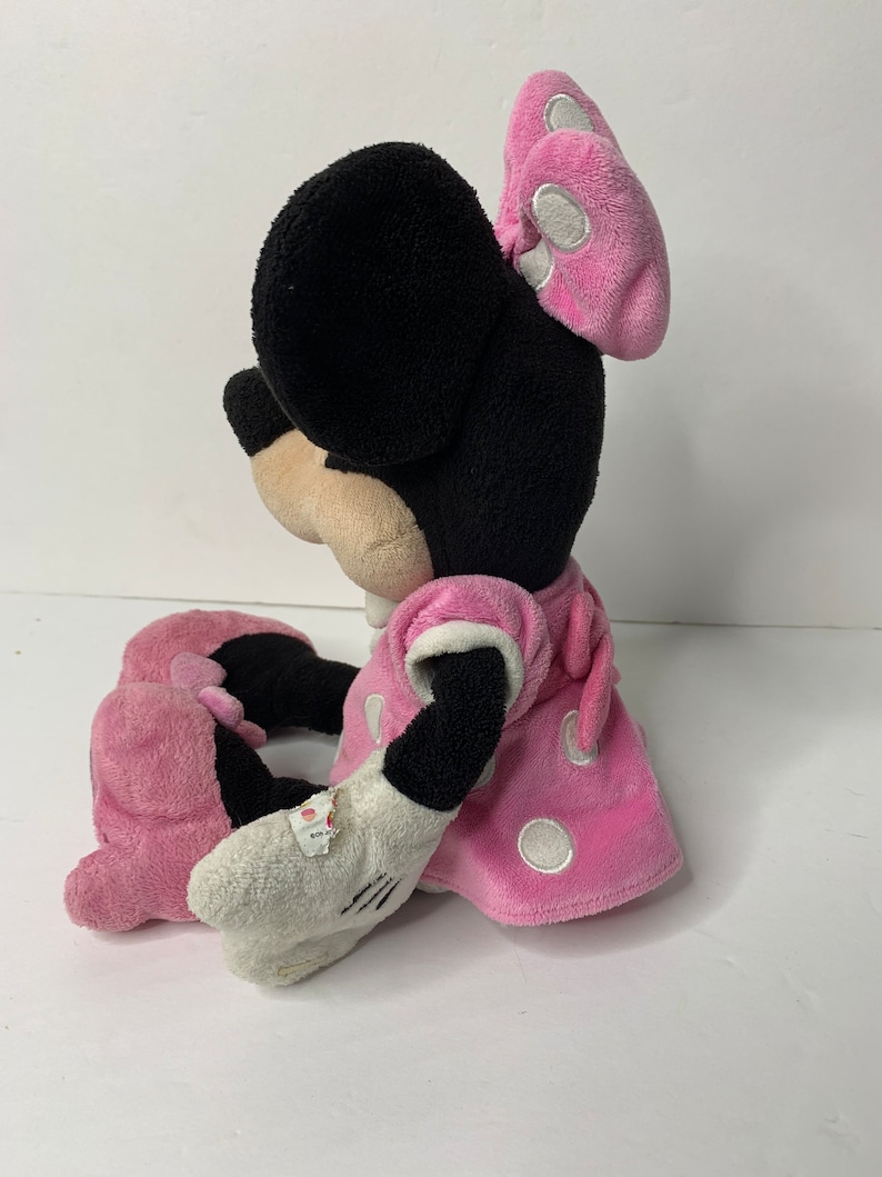 May include: A pink plush Minnie Mouse toy with white polka dots on her dress and bow. She is sitting with her legs crossed and her arms resting on her lap.
