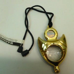 May include: A gold-colored pendant with a black cord. The pendant has a unique shape with a circular opening and a smaller circular design at the top. A tag with the word "WARNING" is attached.