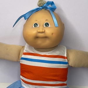 Puede incluir: Una muñeca Cabbage Patch Kids vintage con cabeza calva, ojos verdes y un pequeño lazo azul. La muñeca lleva una camiseta a rayas blancas, naranjas y azules y una falda de animadora azul con ribete dorado. Los brazos de la muñeca están extendidos.