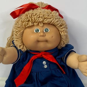 Könnte beinhalten: Eine Puppe mit hellbrauner Haut, grünen Augen und blondem Wollhaar, das mit einem roten Satinband verziert ist. Die Puppe trägt ein marineblaues Kleid mit weißem Kragen, einer roten Krawatte und drei weißen Knöpfen am Vorderteil.