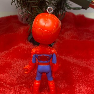 Puede incluir: Adorno navide&ntilde;o de Spider-Man rojo y azul. El adorno es una figura en miniatura del superh&eacute;roe, con cabeza y cuerpo rojos, detalles azules y un lazo plateado para colgar. El adorno est&aacute; sobre un fondo rojo.