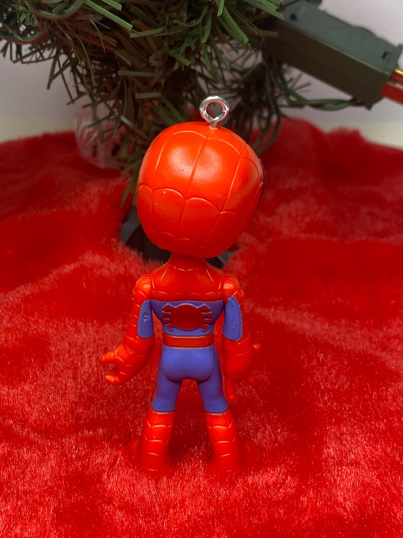 Puede incluir: Adorno navide&ntilde;o de Spider-Man rojo y azul. El adorno es una figura en miniatura del superh&eacute;roe, con cabeza y cuerpo rojos, detalles azules y un lazo plateado para colgar. El adorno est&aacute; sobre un fondo rojo.