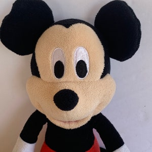 Op de afbeelding: Een Mickey Mouse knuffel. De kop is zwart met grote, afgeronde oren, een beige gezicht en zwarte ogen. Hij draagt een zwart shirt, een rode broek met witte knopen en gele schoenen. Het speelgoed is zacht.