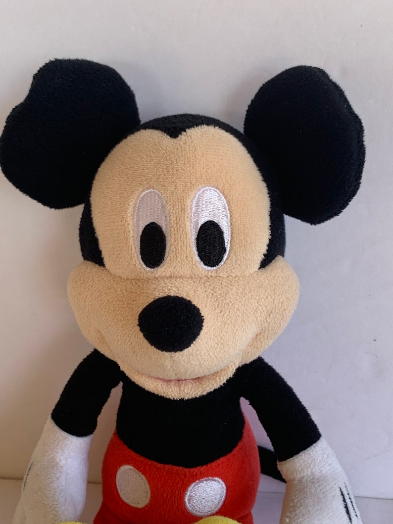 Op de afbeelding: Een Mickey Mouse knuffel. De kop is zwart met grote, afgeronde oren, een beige gezicht en zwarte ogen. Hij draagt een zwart shirt, een rode broek met witte knopen en gele schoenen. Het speelgoed is zacht.