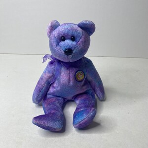 May include: Purple and blue tie-dye beanie baby bear with a black nose and black button eyes. The bear is sitting with a purple ribbon around its neck and a Ty heart tag attached to its chest.