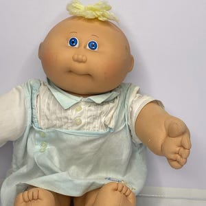 May include: A vintage Cabbage Patch Kid doll with a bald head, blue eyes, and a yellow yarn bow. The doll wears a light blue and white outfit with a ruffled collar and button details. The doll's skin is a peach colour.