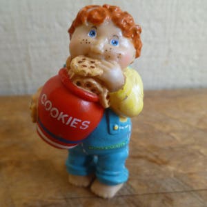 Puede incluir: Una figurita de un tarro de galletas de cerámica roja y azul que representa a un niño con cabello rojo, ojos azules y una galleta en la boca. El tarro dice "COOKIES" en el lateral.