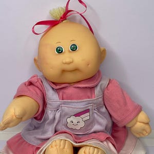 Peut inclure: Poupée Cabbage Patch Kid vintage avec une tête chauve, des yeux verts et une expression souriante. La poupée porte une robe rose avec un tablier blanc et lavande avec un arc-en-ciel et un nuage. Un ruban rose et de la laine jaune ornent le haut de la tête.