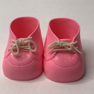 May include: A pair of pink baby shoes with white laces. The shoes have a classic oxford style with decorative perforations on the toe. The shoes are made of a smooth, solid material and are designed for infants.