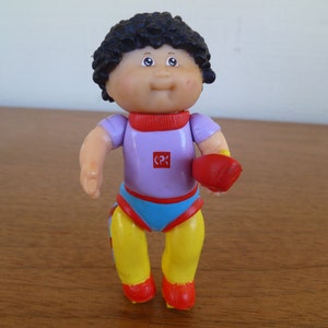 May include: A Cabbage Patch Kid doll wearing a purple and blue outfit with yellow and red roller skates. The doll has black hair and a red heart on its chest.