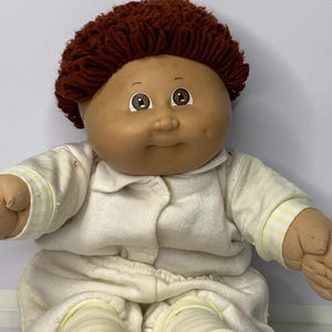 Könnte beinhalten: Eine Vintage-Puppe mit einem Vinylgesicht, braunem Garnhaar und braunen Augen. Die Puppe trägt einen cremefarbenen Strampler mit gelb gestreiften Akzenten. Die Arme und Beine der Puppe sind aus weichem Stoff.
