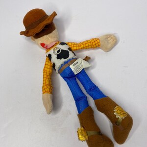 May include: A plush toy of the character Woody from the Disney Pixar film Toy Story. The doll features a brown cowboy hat, a yellow plaid shirt, blue pants, and brown boots. The vest is white with a black cow pattern.
