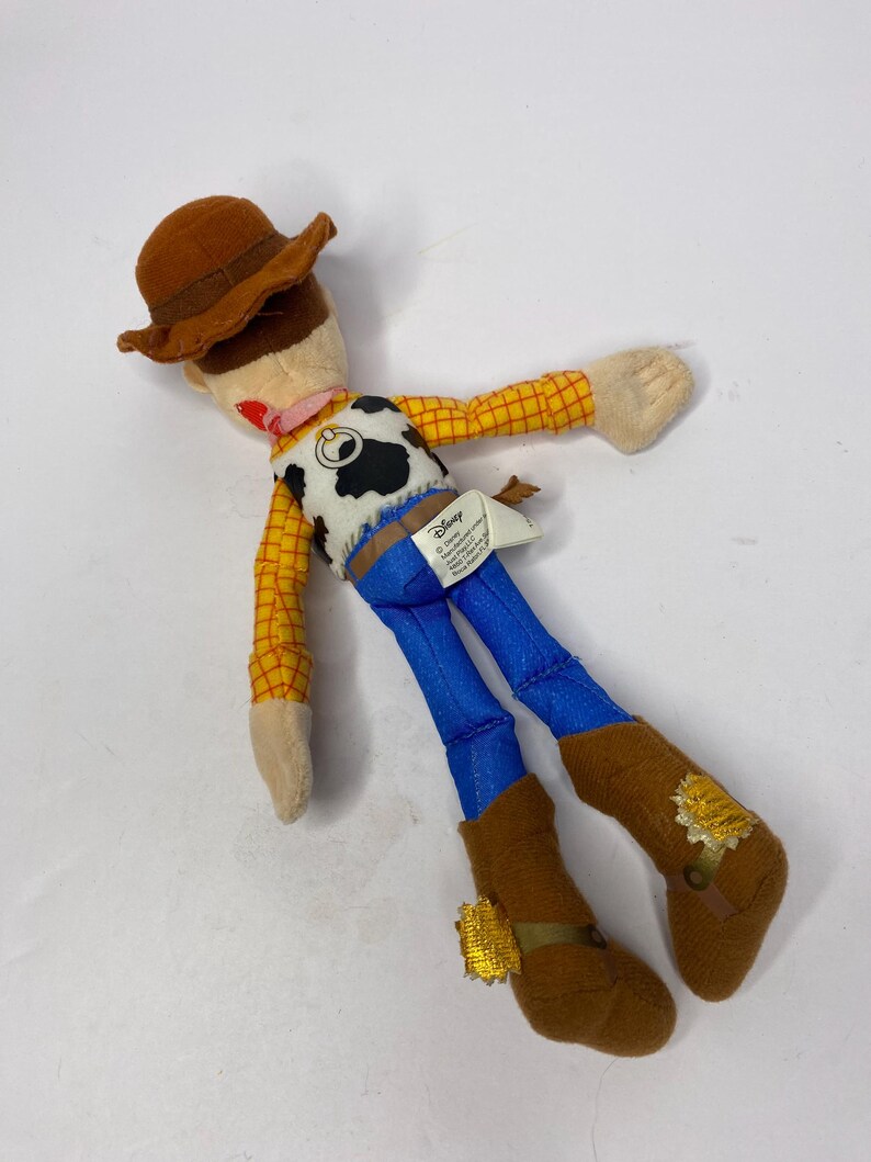 May include: A plush toy of the character Woody from the Disney Pixar film Toy Story. The doll features a brown cowboy hat, a yellow plaid shirt, blue pants, and brown boots. The vest is white with a black cow pattern.