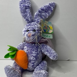May include: A plush lavender bunny with long ears and a pink nose, holding an orange carrot with green leaves. The bunny has a tag that says "Fiesta". A soft, decorative item for Easter.