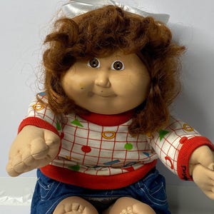 Può includere: Una bambola Cabbage Patch Kid con capelli castani ricci, che indossa un fiocco bianco, una camicia a motivi geometrici rossi e bianchi e una gonna di jeans blu.