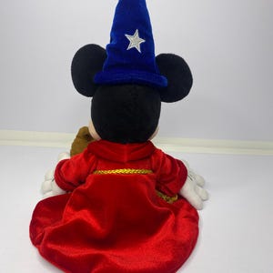 May include: A plush Mickey Mouse dressed as a sorcerer, viewed from the back. The figure wears a red velvet robe with gold trim and a blue velvet wizard hat with a silver star. The plush has black ears and white gloves.