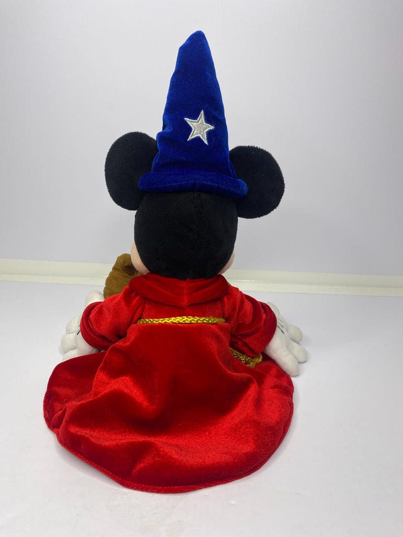 May include: A plush Mickey Mouse dressed as a sorcerer, viewed from the back. The figure wears a red velvet robe with gold trim and a blue velvet wizard hat with a silver star. The plush has black ears and white gloves.