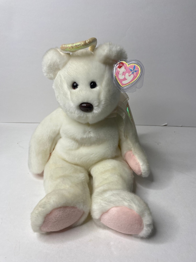 May include: White plush angel bear with a halo and pink feet. The bear has a brown nose and black eyes. The bear is sitting on a white surface. The bear has a Ty tag attached to its ear.