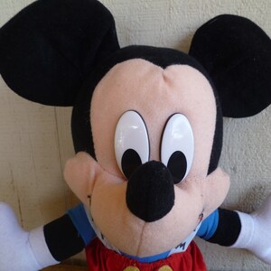 Może przedstawiać: Pluszowa zabawka Mickey Mouse z czarnymi uszami, czarnym nosem i dużymi białymi oczami. Zabawka ma na sobie czerwony i niebieski str&oacute;j.
