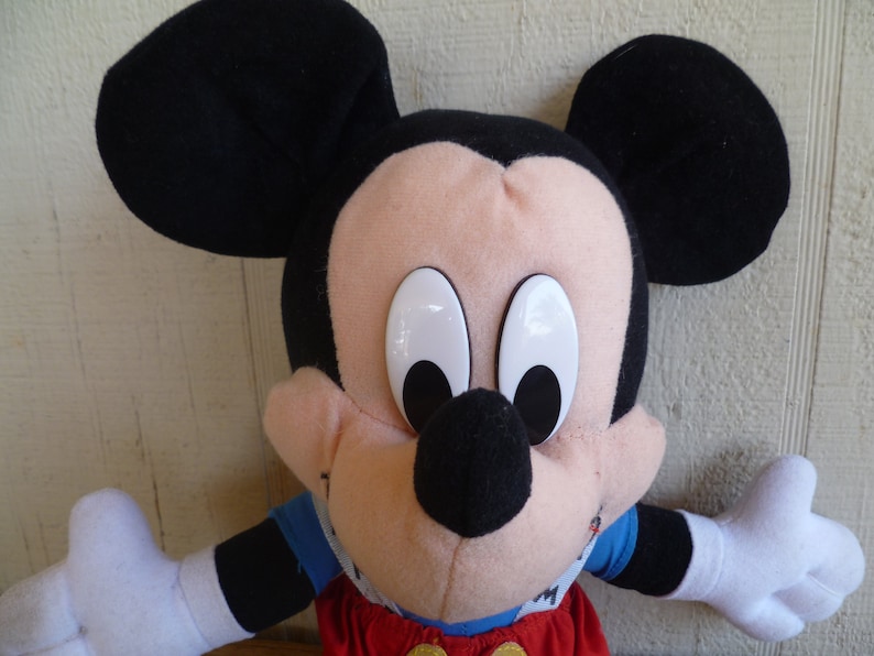 Może przedstawiać: Pluszowa zabawka Mickey Mouse z czarnymi uszami, czarnym nosem i dużymi białymi oczami. Zabawka ma na sobie czerwony i niebieski str&oacute;j.