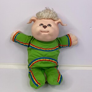 May include: A plush doll with a soft, light-toned face, brown eyes, and blonde hair. The doll wears a bright green long-sleeved jumpsuit featuring horizontal rainbow-colored stripes across the chest, sleeves, and legs, with a small brown nose.