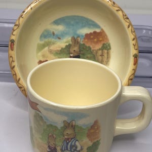 May include: A cream-colored ceramic cup and bowl set featuring illustrations of rabbits in a pastoral scene. The bowl has a decorative border with carrots. The cup and bowl are likely part of a children's dish set.