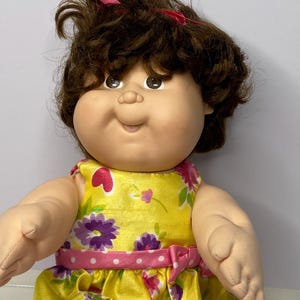 Könnte beinhalten: Eine Vintage-Puppe mit braunen Locken und braunen Augen. Die Puppe trägt ein gelbes Kleid mit Blumenmuster und einen rosa gepunkteten Gürtel. Die Arme und Beine der Puppe sind aus weichem Stoff.