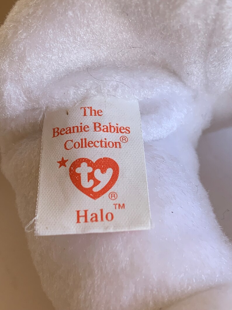 May include: White Beanie Baby plush toy with a red tag that reads "The Beanie Babies Collection&reg;" with a star and the Ty logo. The tag also reads "Halo" with a registered trademark symbol.