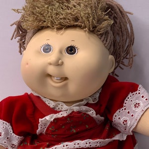 Könnte beinhalten: Eine Cabbage Patch Kid Puppe mit braunen Garnhaaren, die ein rotes Kleid mit weißen Spitzenbesatz und einem grün-roten Blumenmuster trägt.