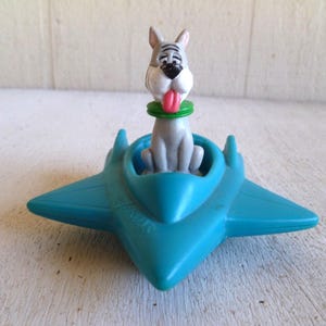 May include: A vintage toy featuring a cartoon dog figure with a pink tongue and green collar, seated in a teal-colored airplane. The toy is made of plastic and has a retro aesthetic.