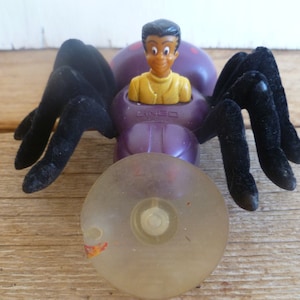 May include: A purple and black toy spider with a suction cup on the bottom. A cartoon figure with brown hair and a yellow shirt is sitting in the spider's body. The figure is labeled "Bingo".