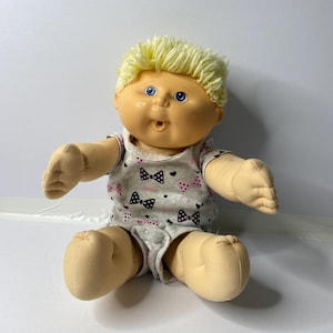 Puede incluir: Una muñeca bebé vintage con cabello rubio de hilo y ojos azules. La muñeca lleva un body gris con un estampado de lazos rosas y negros. La muñeca está sentada con los brazos y las piernas extendidos.