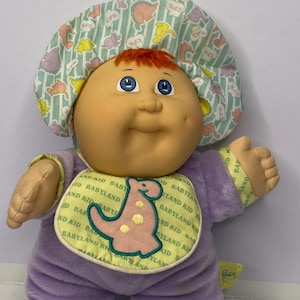 May include: A vintage Cabbage Patch Kid doll with a round, peach-coloured face, blue eyes, and red hair. It wears a lavender onesie, a bib with a dinosaur design, and a patterned bonnet. The doll is a soft plush toy.