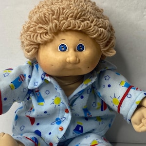 May include: A Cabbage Patch Kid doll with blonde yarn hair wearing blue pajamas with a rocket ship and star pattern. The doll has blue eyes and a rosy complexion.