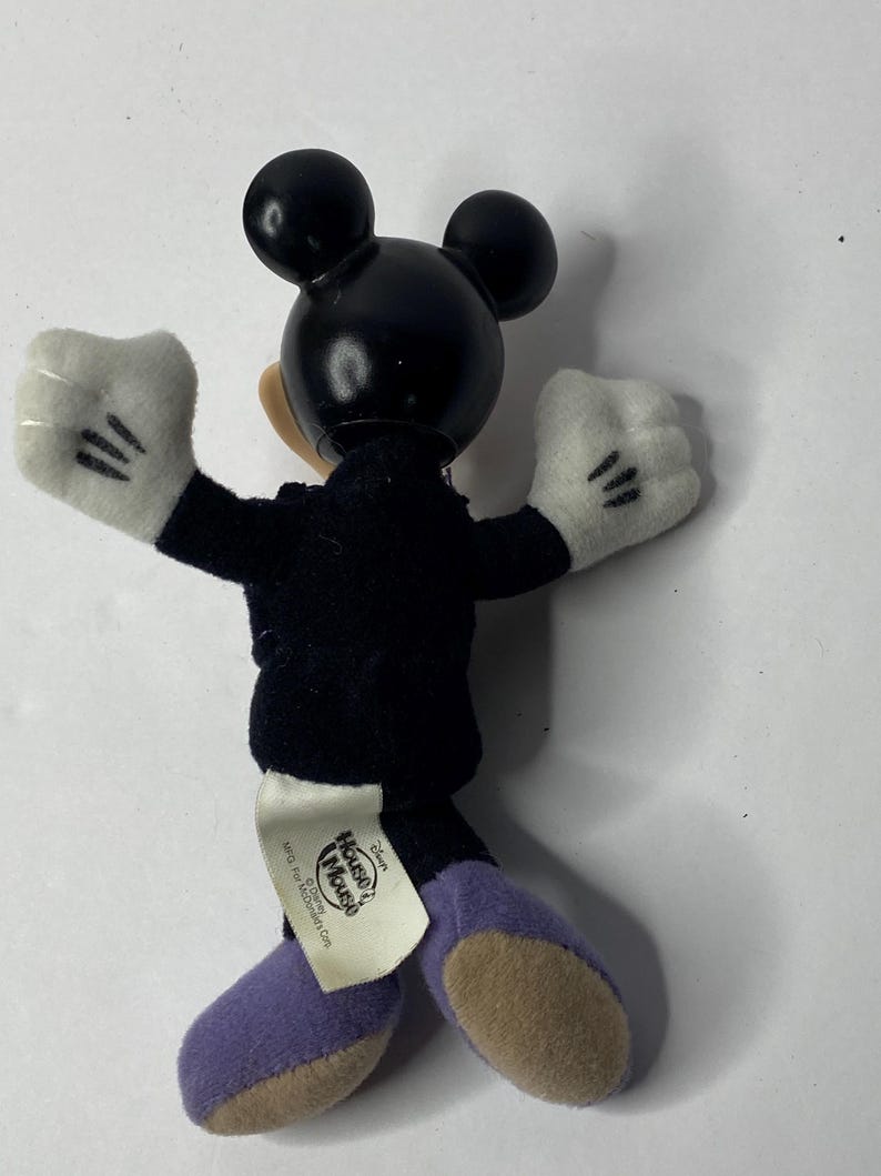 May include: A black and white plush Mickey Mouse toy wearing a black suit and purple shoes. The toy has a tag on its back that says "Disney's House of Mouse" and "&copy; Disney.  All rights reserved."