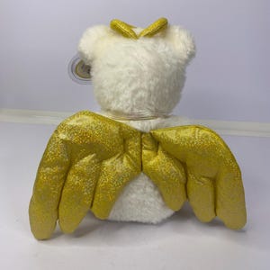 K&ouml;nnte beinhalten: Ein wei&szlig;es Pl&uuml;schtier mit goldglitzernden Fl&uuml;geln und einem goldenen Heiligenschein. Das Spielzeug hat ein angebrachtes Etikett. Die Fl&uuml;gel sind ausgebreitet und der Heiligenschein sitzt auf dem Kopf des Spielzeugs. Das Spielzeug befindet sich auf wei&szlig;em Hintergrund.