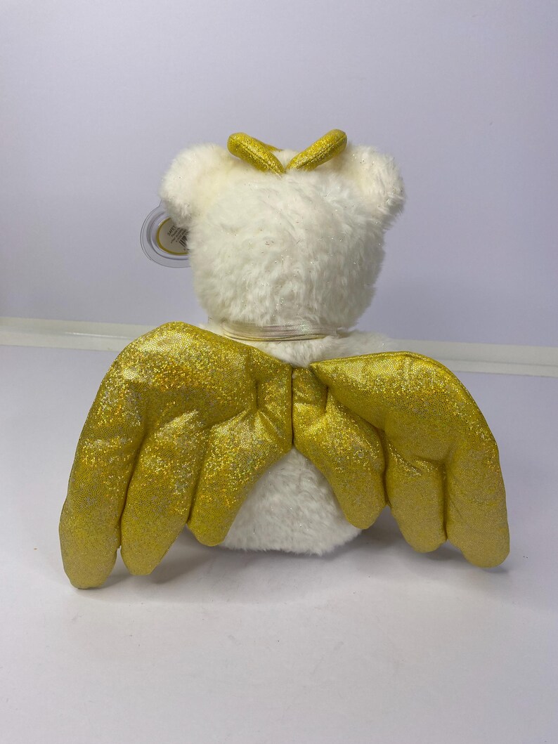 K&ouml;nnte beinhalten: Ein wei&szlig;es Pl&uuml;schtier mit goldglitzernden Fl&uuml;geln und einem goldenen Heiligenschein. Das Spielzeug hat ein angebrachtes Etikett. Die Fl&uuml;gel sind ausgebreitet und der Heiligenschein sitzt auf dem Kopf des Spielzeugs. Das Spielzeug befindet sich auf wei&szlig;em Hintergrund.