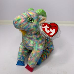 May include: A colourful plush goat toy with green eyes, green horns, and a yellow beard. The goat has a rainbow pattern and a red heart-shaped tag that says "Ty". The toy is sitting on a white surface.