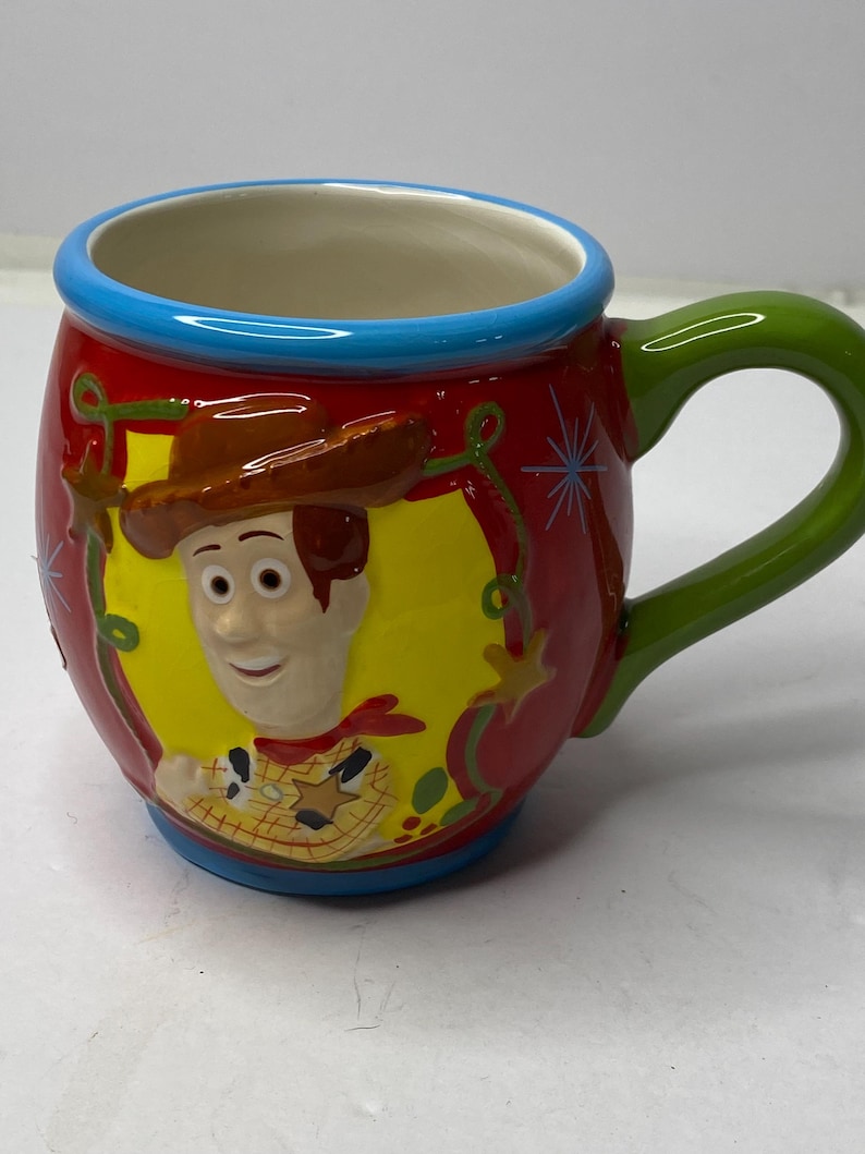 Puede incluir: Una taza de cer&aacute;mica con una imagen de dibujos animados de Woody de Toy Story. La taza tiene un exterior rojo con un marco ovalado amarillo alrededor de la cara de Woody. El asa es verde y el borde es azul.