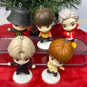 May include: Five small, cartoon figurines with large heads and stylized features. Each figure has a different hairstyle and outfit, including a black bucket hat and a red jacket. They are standing on white circular bases, with a Christmas tree in the background.