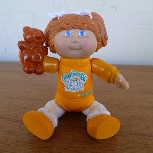 Op de afbeelding: Een Cabbage Patch Kids pop die een oranje shirt draagt met het Cabbage Patch Kids logo. De pop heeft rood haar en blauwe ogen en houdt een bruine teddybeer vast.