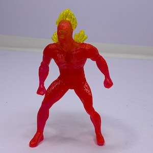 May include: A red and yellow action figure of a superhero in a standing pose. The figure has a flame-like design on the head and shoulders, with a red body and a gradient from red to pink on the legs and arms.