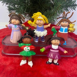 May include: Five vintage Cabbage Patch Kids Christmas ornaments, each with a unique outfit and hairstyle. One doll plays a trumpet, another holds a star wand, and another a teddy bear. The ornaments are on a red, fuzzy surface with a green Christmas tree.