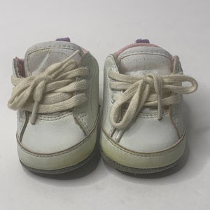 Könnte beinhalten: Ein Paar weiße Lederschuhe für Babys mit weißen Schnürsenkeln und einem violetten und rosa Besatz um den Knöchel.