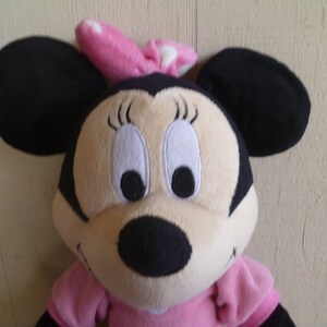 May include: A plush toy of Minnie Mouse wearing a pink bow with white polka dots. She has black ears, a black nose, and large white eyes with black pupils. She is wearing a pink dress.