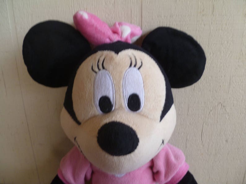 May include: A plush toy of Minnie Mouse wearing a pink bow with white polka dots. She has black ears, a black nose, and large white eyes with black pupils. She is wearing a pink dress.