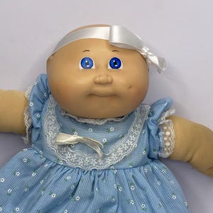 Pode incluir: Uma boneca vintage com cabeça careca, olhos azuis e tom de pele claro. Ela usa um vestido azul claro com estampas florais brancas e acabamento em renda. Uma fita branca é amarrada em volta da cabeça e um laço no decote.