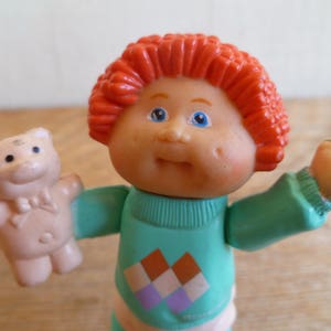 Puede incluir: Una muñeca de pelo rojo con un suéter azul y verde con un patrón de diamantes. La muñeca sostiene un oso de peluche rosa con una corbata de lazo.