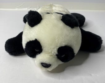 May include: A black and white plush panda bear laying on its belly with its head turned to the side. The panda has black eyes and a black nose.